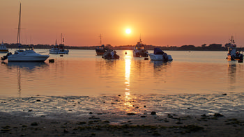 Sunset Christchurch harbour This landscape photograph captures an evening sunset over Christchurch Harbour in Dorset, United Kingdom. The warm light of the sun, low in the sky, reflects across the tranquil water, marking a late spring day. Several boats are anchored near the beach, emphasising the harbour’s role in local transport and maritime activity. The image features the natural beauty of Christchurch, with the boats silhouetted against the colourful sky and the shoreline in the foreground.
